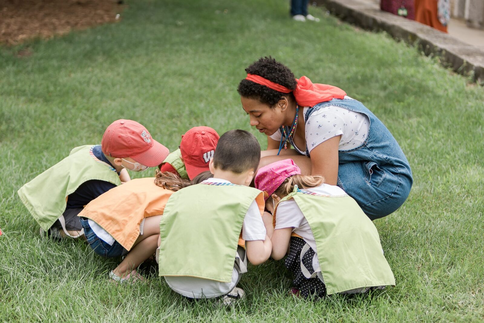 A teacher crouching down with a group of young children gathered in a circle on the grass.