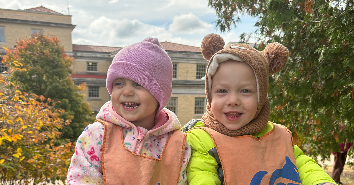 Two children smile sitting outdoors in the fall with stocking hats on.