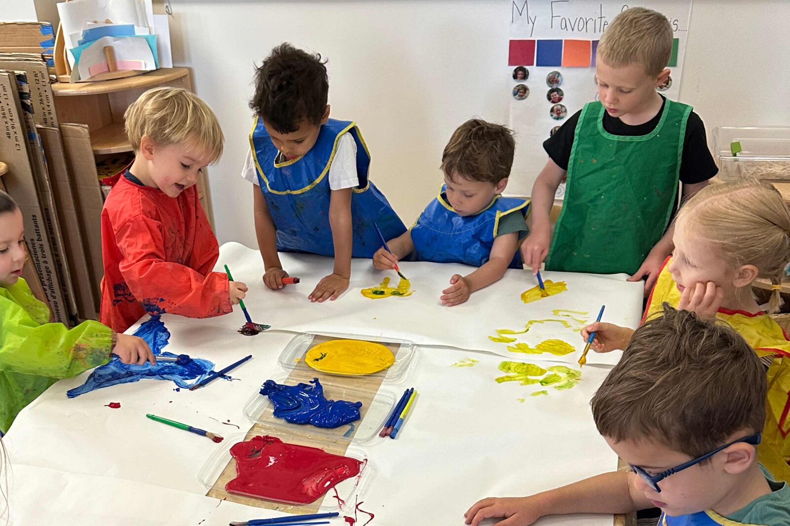 Seven kids sit around a table painting with red, blue and yellow.